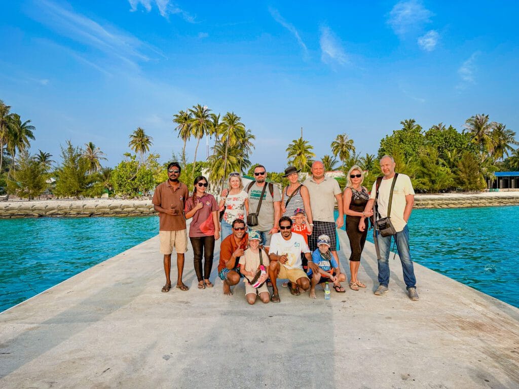 A group picture of our guests and employees taking an arrival photo at Fodhdhoo jetty, with the island's lush greenery in the background.