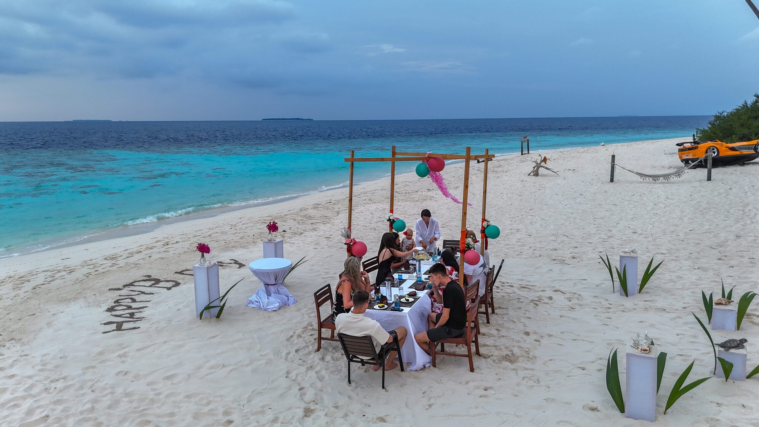 Group of friends sharing sunset tea on the beach while pastel clouds reflect on calm water