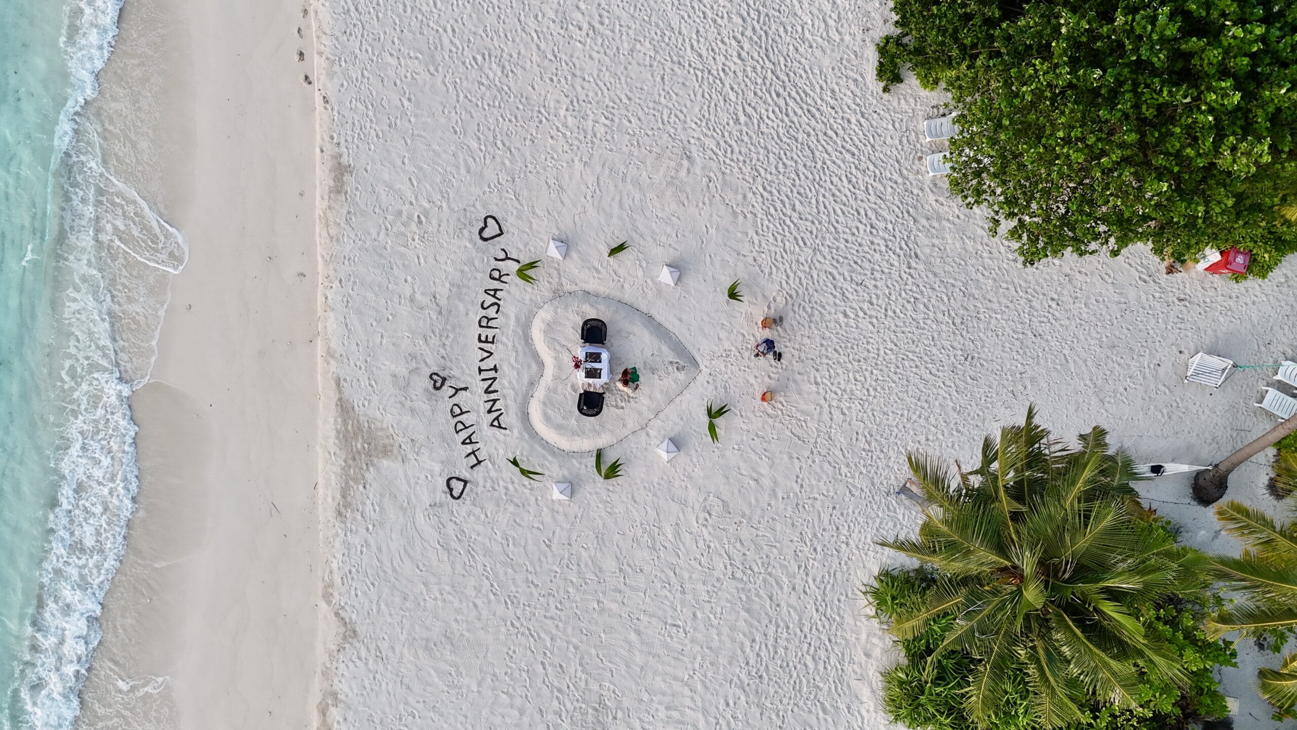 Aerial image of romantic anniversary dinner setup with lanterns and rose petals beside gentle surf