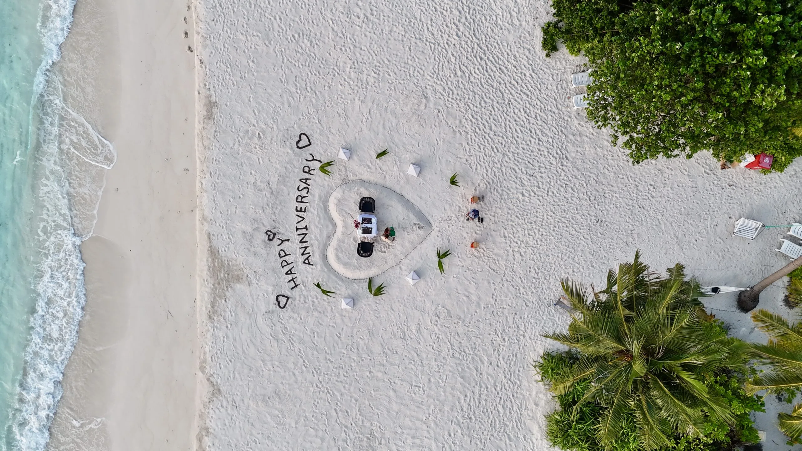 Aerial image of romantic anniversary dinner setup with lanterns and rose petals beside gentle surf